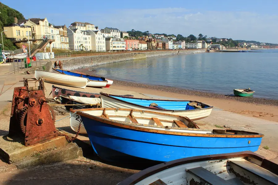 Painted seafront houses above the railway sea wall curving round a red-sand South Devon beach, with rowing boats drawn up in the foreground