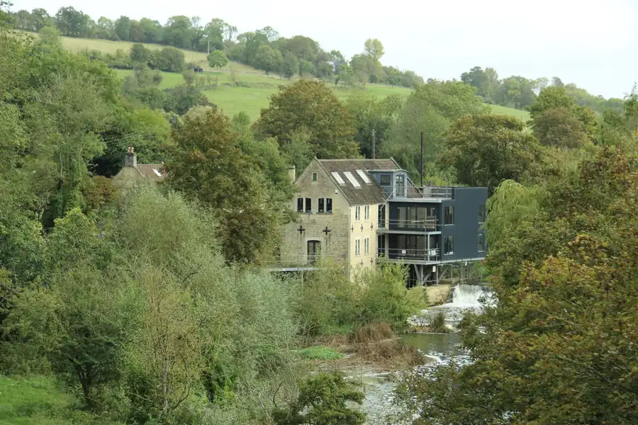 Stone mill with a modern dark glazed extension beside a weir, the kind of conversion where large steel-look glazing is priced